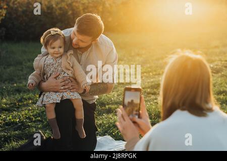 Glückliche junge Familie, die im Freien Picknick macht, Frau, die Fotos von ihrem Mann und ihrer kleinen Tochter bei Sonnenuntergang macht Stockfoto