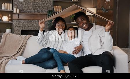 Afrikanisches Paar sitzt auf der Couch unter dem Kartondach lächelnd und schaut auf die Kamera aktiv glücklich Tochter Mädchen läuft sitzt auf dem Sofa. Umzugstag zu Neu Stockfoto