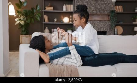 Afroamerikanische liebevolle Mutter Mama, die auf dem Sofa liegt und sich im Wohnzimmer entspannt, mit kleinen Töchtern, die sich unterhalten und Spaß haben, zwangloses Spiel spielen, halten Stockfoto