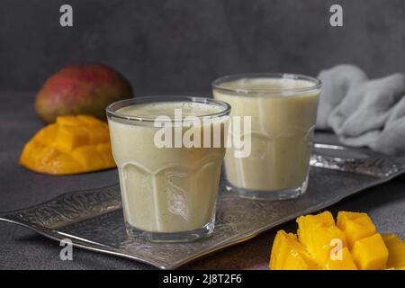 Mango-Lassi in zwei Gläsern auf grauem Hintergrund. Frische indischer Lassi aus Joghurt, Gewürzen, Mango und Eis Stockfoto