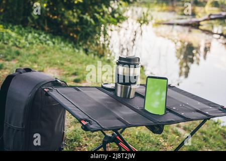 Campingtisch mit Thermobecher und Smartphone im Freien. Grüner Bildschirm. Malerische Landschaft. Freizeit- und Reisekonzept. Technologie Stockfoto