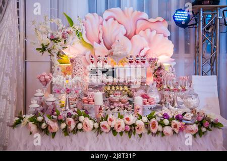 Köstliche Desserts an der Hochzeitstheke im Buffetbereich: Verzierte Kerzen, Rosenknospen, Bänder, Makronen, Erdbeeren in weißer Schokolade. Stockfoto