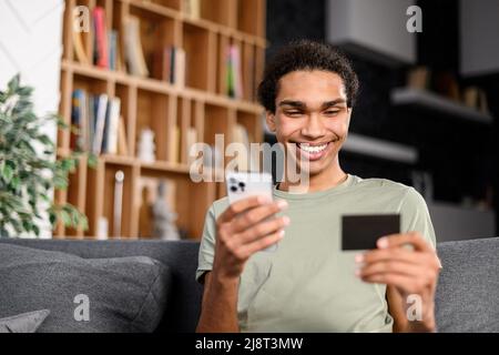 Schöner Mann macht online kaufen. Ein Hipster trägt ein legeres Hemd und hält ein Handy und eine Kreditkarte bestellt Essen. Lächelnder Kerl verwendet Smartphone und EC-Bankkarte für die Zahlung im E-Shop Stockfoto