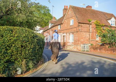 Rückansicht des Mannes, der auf einer schmalen Straße durch ein typisch englisches Landdorf bei Herbstsonne reitet. Stockfoto