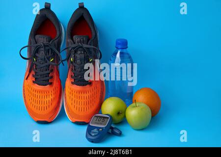 Orangefarbene Turnschuhe, grüne Äpfel, Orangen, eine Flasche Wasser und Blutzuckermessgerät auf blauem Hintergrund mit Kopierfläche. Therapie gegen Insulinresistan Stockfoto