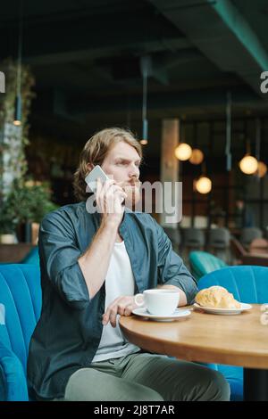 Seriös schöner junger Mann mit Bart, der am Tisch sitzt und einen Kollegen telefonisch anruft, während er im Café auf ihn wartet Stockfoto