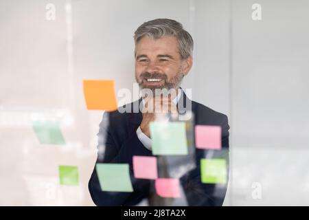 Brainstorming. Lächelnder, reifer Geschäftsmann, der auf Haftnotizen an der transparenten Glaswand schaut Stockfoto