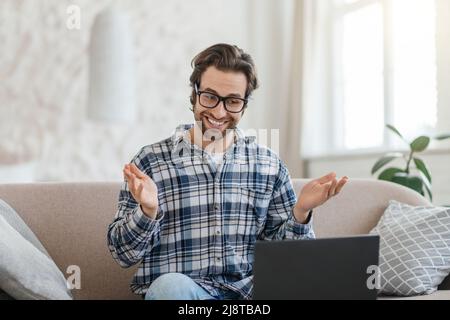 Der fröhliche junge kaukasische Lehrer mit Stoppeln in der Brille sitzt auf dem Sofa mit Laptop und gestikuliert Stockfoto