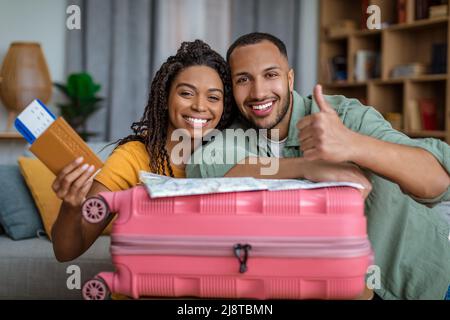 Urlaubskonzept. Aufgeregte afroamerikanische Ehegatten halten Pässe und zeigen den Daumen hoch, lehnen sich auf den Koffer Stockfoto