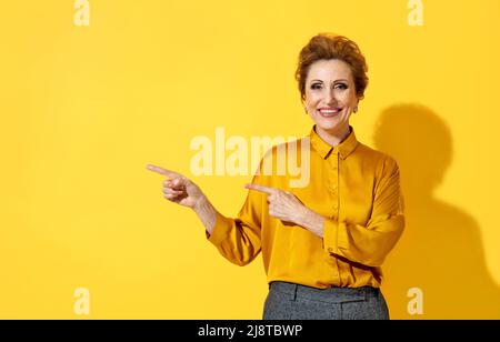 Lächelnde Frau zeigt mit beiden Zeigefinger weg, zeigt auf dem Kopierplatz für Ihren Text. Foto einer attraktiven älteren Frau in gelbem Hemd auf gelbem Rücken Stockfoto