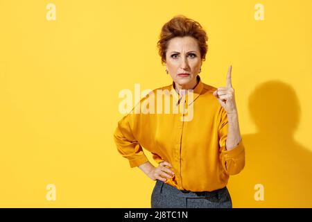 Ältere, strenge Frau zeigt mit dem Zeigefinger nach oben, warnt, benachrichtigt, empfiehlt etwas. Foto von attraktiven Frau in gelbem Hemd auf gelbem Hintergrund Stockfoto