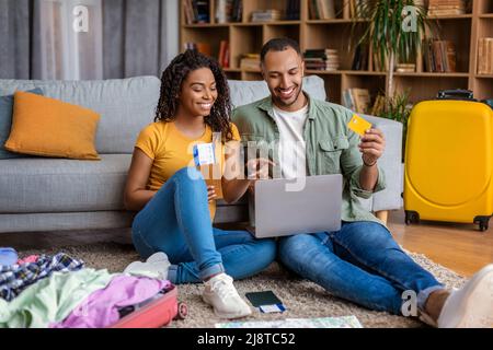 Aufgeregte schwarze Ehepartner, die ihren Laptop benutzen und das Hotel online mit Kreditkarte buchen, sitzen zu Hause mit Koffern auf dem Boden Stockfoto