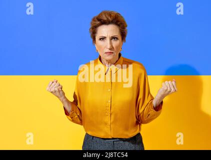 Ukrainische Frau, die vor Wut die Fäuste zusammenhält, bereit zu kämpfen. Foto einer attraktiven Frau in gelbem Hemd vor dem Hintergrund der Flagge der Ukraine Stockfoto