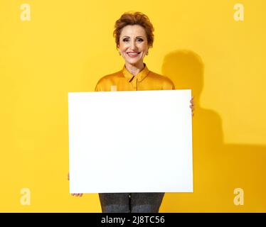 Lächelnde Frau mit weißem leerem Brett. Foto von attraktiven älteren Frau in gelbem Hemd auf gelbem Hintergrund Stockfoto