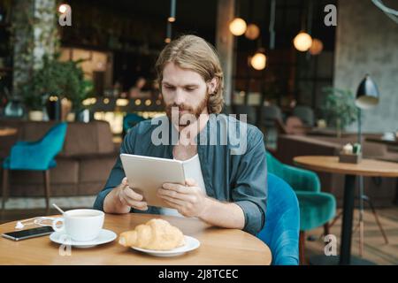 Konzentrierter junger bärtiger Mann im lässigen Hemd, der Kaffee trinkt und Croissant isst, während er im Café sein Smartphone überprüft Stockfoto