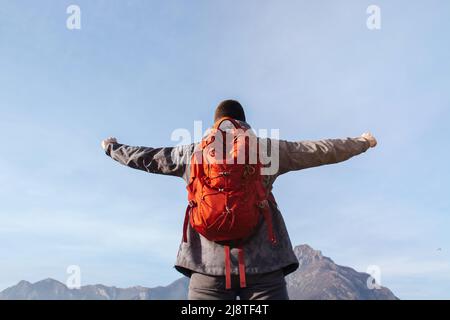 Rückansicht der Reisende des Mannes genießt die Freiheit mit den aufgehobenen Armen und dem roten Touristenrucksack, der bei Sonnenuntergang auf dem Berg steht. Blauer Himmel. Konzept der tou Stockfoto