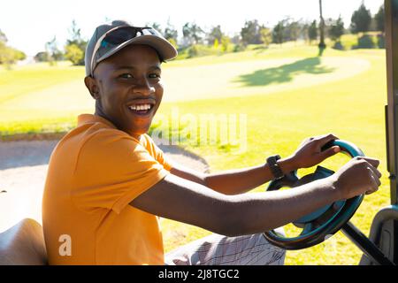 Porträt eines glücklichen afroamerikanischen jungen Mannes mit Mütze, der auf dem Golfplatz einen Golfwagen fährt Stockfoto