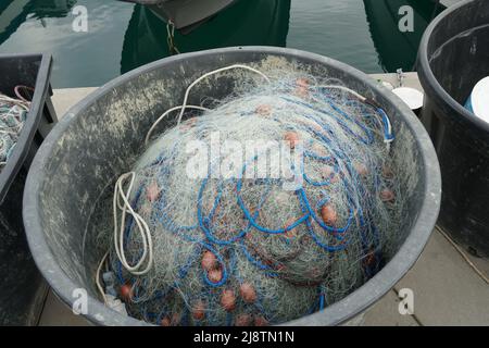 Fischernetz mit blauen Seilen und rot-braunen Schwimmern in schwarzem ovalem Plastikeimer gelagert, bereit zum Laden auf Fischerboot. Stockfoto