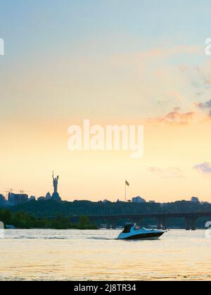 Blick auf den Sonnenuntergang über dem Mutterland-Denkmal, Motorboot auf dem Dnipro-Fluss, Paton-Brücke. Kiew, Ukraine Stockfoto