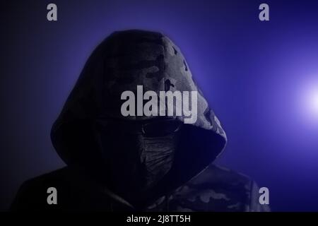 Stilvoller Mann in dunklem Hoodie, schwarzer Brille und dunkelem Hintergrund in blauem Licht, Hacker, Angreifer und Bandit Stockfoto