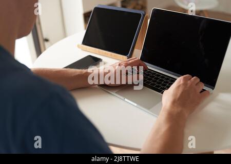 Eine seriöse Person, die zu Hause aus der Ferne arbeitet. Ein Mann gibt Informationen in einen Laptop ein. Layout-Konzept, Design. Schwarze Bildschirme auf Laptops und Tablets. Layout für Ihr Design. Hochwertige Fotos Stockfoto