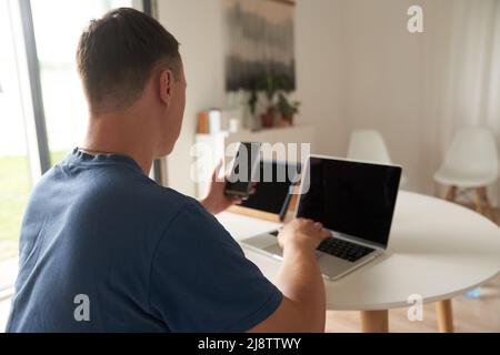 Eine seriöse Person, die zu Hause aus der Ferne arbeitet. Betrachtet das Telefon und gibt Informationen im Laptop ein. Hochwertige Fotos Stockfoto