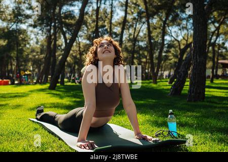 Aktive junge Frau in Sportkleidung, die im Stadtpark im Freien Yoga praktiziert. Auf Fitnessmatte legen und ihren Körper strecken, nach unten gerichtete Yoga-Haltung des Hundes, Stockfoto