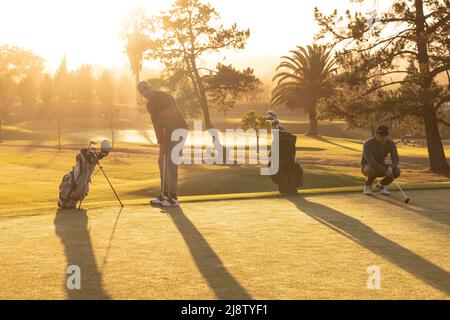 Multirassische junge männliche Freunde, die auf dem grasbewachsenen Land gegen Bäume und am Himmel auf dem Golfplatz Golf spielen Stockfoto
