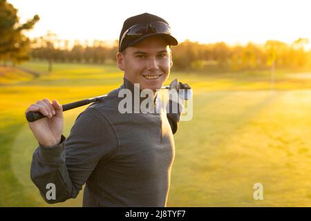 Porträt eines lächelnden kaukasischen jungen Mannes mit Mütze, der den Golfschläger auf dem Golfplatz hält Stockfoto