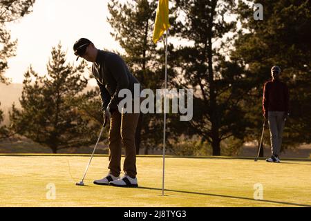 Kaukasischer junger Mann spielt Golf mit einem afroamerikanischen jungen Freund auf dem Golfplatz bei Sonnenuntergang Stockfoto