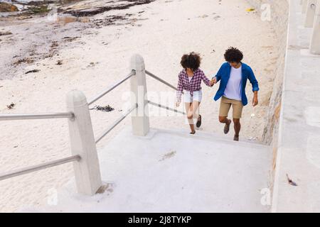 Liebevolles junges afroamerikanisches Paar, das sich die Hände hält, während es am Strand Stufen klettert Stockfoto