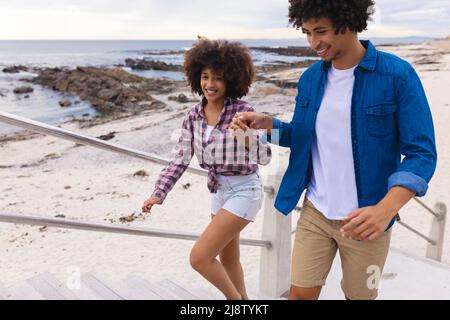 Lächelndes, junges afroamerikanisches Afropaar hält sich die Hände, während es am Strand Stufen klettert Stockfoto