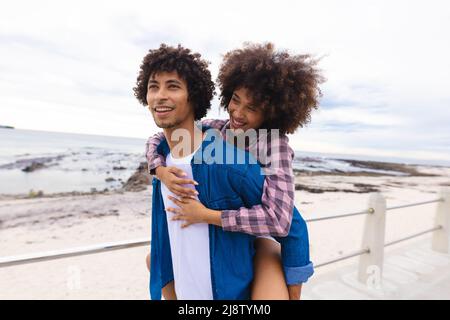 Lächelnder junger Mann Huckepack afro afro afroamerikanische Freundin am Strand, kopieren Raum Stockfoto