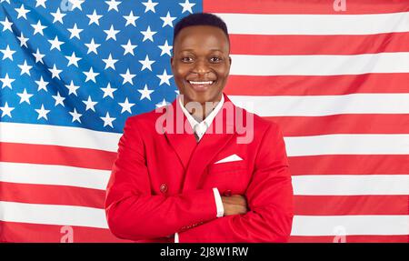 Glücklicher, gutaussehender afroamerikanischer Mann in rotem Anzug, der vor dem Hintergrund der US-Flagge steht Stockfoto