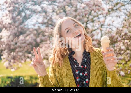 Schöne junge blonde 20s Mädchen hält cremiges Eis in Waffelkegel und zeigt Friedenszeichen im Stadtpark. Blühende rosa Blüten Magnolie Hintergrund Stockfoto