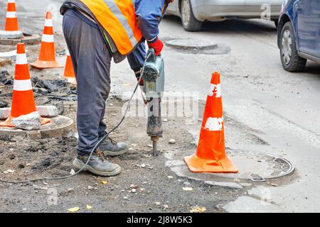 Ein Straßenarbeiter arbeitet mit einem elektrischen Presslufthammer auf einem Straßenabschnitt, der mit Verkehrskegeln eingezäunt ist. Stockfoto