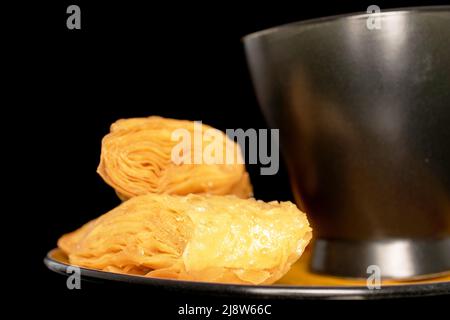 Zwei süße Honigbaklava auf einer Untertasse mit schwarzer Tasse, Nahaufnahme, isoliert auf schwarzem Hintergrund. Stockfoto