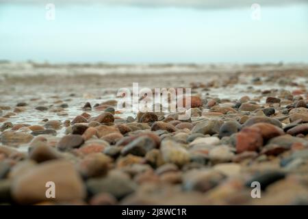 Steine liegen wunderschön an der Küste. Steine auf dem Sand. Viele bunte Muscheln und Steine liegen am Strand an einem sonnigen Tag mit blauem Himmel und einem Stockfoto