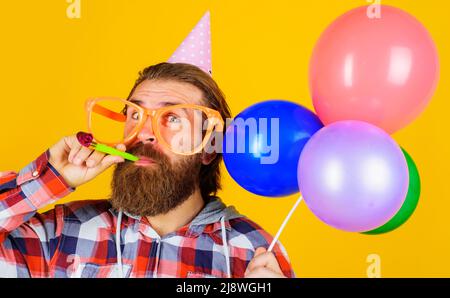 Bärtiger Mann in Partyhut und Brille mit Luftballons, die Noisemaker blasen. Geburtstagsfeier. Stockfoto