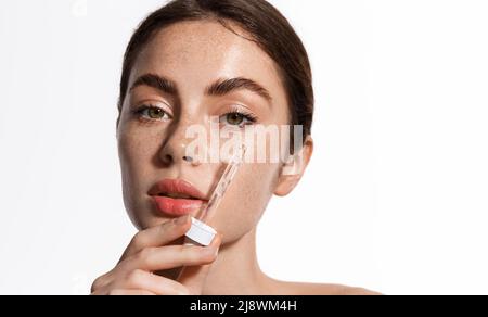 Kopfporträt der Frau mit dicken natürlichen Augenbrauen, Sommersprossen, saubere Haut mit Feuchtigkeit nach der Gesichtscreme, hält Tropf mit Kollagen, Vitamin c Stockfoto