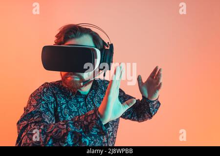Millenialer bärtiger Kaukasianer, der ein Virtual-Reality-Headset trägt und ein spiel aus dem jahr 3D spielt. Studio auf orangefarbenem oder rotem Hintergrund aufgenommen. . Hochwertige Fotos Stockfoto