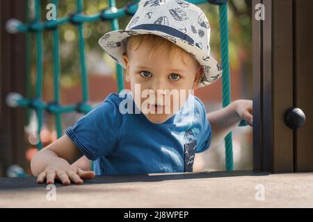 Netter kleiner Junge, der im Sommer auf dem Spielplatz spielt. Ein Porträt eines Kindes in einem Hut blickt mit einem überraschenden Gesichtsausdruck direkt in die Kamera Stockfoto