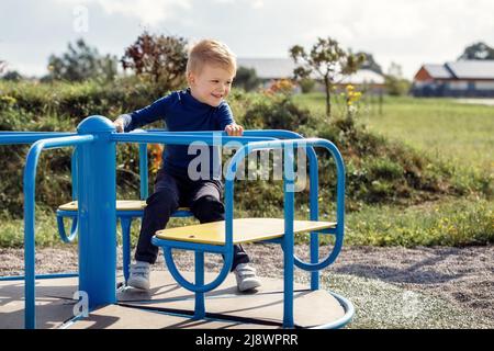 Kaukasischer Junge dreht sich auf einem Kinderkarussell auf einem Spaziergebiet für Kinder in der Stadt. Der Junge ist glücklich und lächelt. Stockfoto
