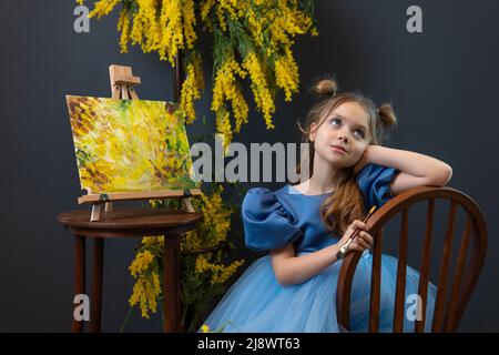 Ein Mädchen sitzt in der Nähe einer Mimose und malt ein Bild in ein blaues Kleid Mimosa Pinsel kleines Mädchen, für Porträt-Zöpfe aus Blumen von Lächeln kaukasischen, Kind Stockfoto