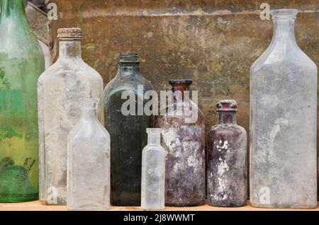 Nahaufnahme von alten, ausgegrasten Alkohol- und Medikamentenglasflaschen vor einem verrosteten Metallbehälter-Hintergrund. Stockfoto