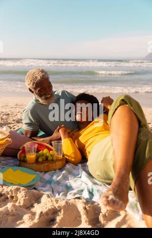 afroamerikanische reife Frau, die auf dem Schoß eines älteren Mannes bei Essen und Trinken liegt, gegen Meer und klaren Himmel Stockfoto