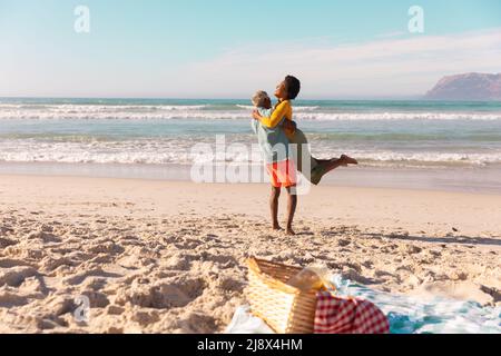 Glücklicher afroamerikanischer älterer Mann, der eine reife Frau trägt, während er am Strand gegen Meer und Himmel steht Stockfoto