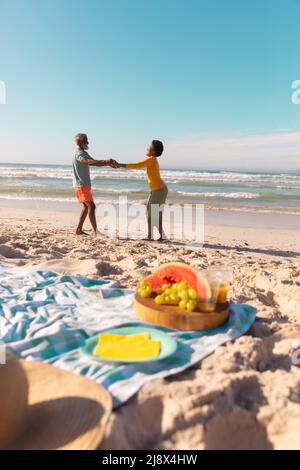 Seitenansicht eines afroamerikanischen Paares, das die Hände hält und am Sandstrand vor blauem Himmel tanzt Stockfoto