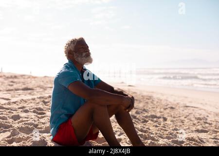 Nachdenklicher bärtiger afroamerikanischer Senioren mit grauen Haaren, der am Sandstrand gegen den Himmel sitzt Stockfoto