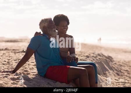 afroamerikanischer bärtiger älterer Mann und reife Frau mit afrohaarigen Haaren sitzen am Strand gegen den Himmel Stockfoto